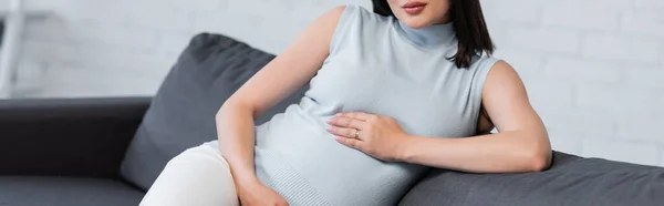 Abgeschnittene Ansicht einer schwangeren Frau, die ihren Bauch berührt, während sie zu Hause auf der Couch sitzt, Banner — Stockfoto