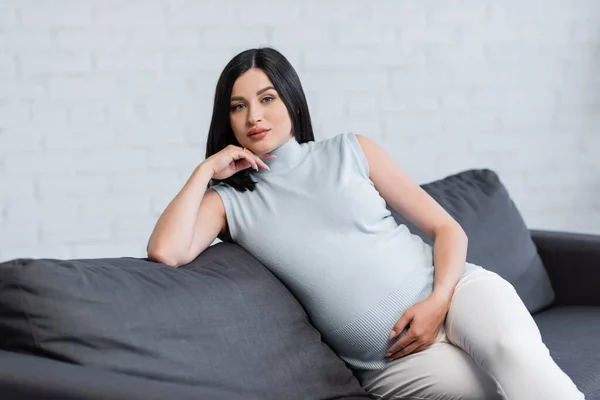 Brunette femme enceinte regardant la caméra tout en étant assis sur le canapé à la maison — Photo de stock