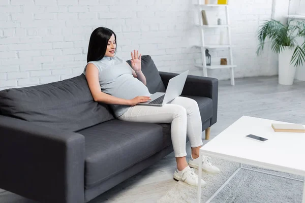 Giovane donna incinta che agita la mano durante la videochiamata sul divano a casa — Foto stock