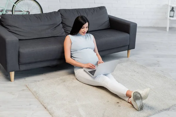 Young pregnant woman sitting on floor near couch and typing on laptop — Stock Photo