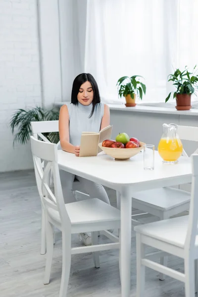 Junge brünette Frau liest Buch in Küche neben reifen Äpfeln und frischem Orangensaft — Stockfoto