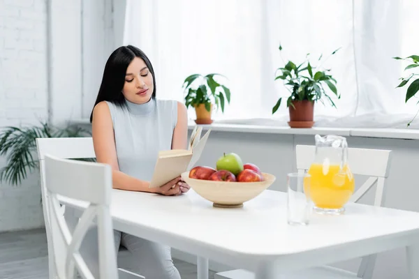 Morena mujer leyendo libro en cocina cerca de manzanas frescas y zumo de naranja en la mesa - foto de stock