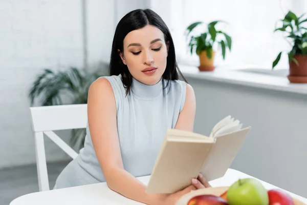 Молода жінка читає книгу біля розмитих яблук на кухонному столі — стокове фото
