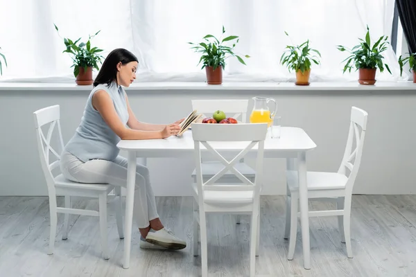 Vista lateral de la mujer embarazada leyendo libro cerca de jugo de naranja y manzanas frescas en la mesa en la cocina - foto de stock