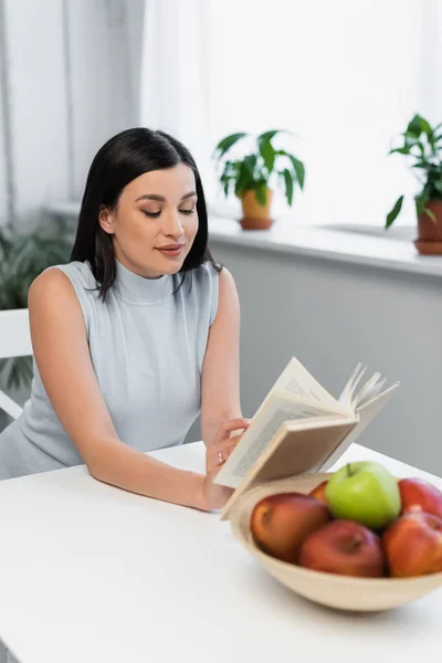 Mujer sonriente leyendo novela cerca de manzanas frescas en la mesa de la cocina - foto de stock