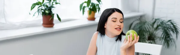 Joven morena sosteniendo manzana fresca cerca de macetas borrosas en la cocina, pancarta - foto de stock