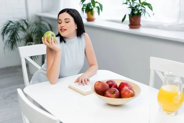 Giovane donna con mela fresca seduta vicino al libro e succo d'arancia in cucina — Foto stock