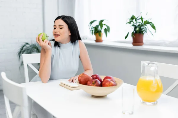 Счастливая брюнетка женщина держит спелое яблоко рядом с книгой и апельсиновый сок на кухонном столе — стоковое фото