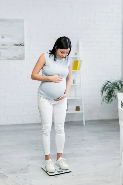 Vista a tutta lunghezza della donna incinta che misura il peso corporeo su scale a pavimento a casa — Foto stock