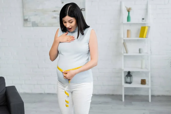 Étonné enceinte toucher la poitrine tout en mesurant le ventre à la maison — Photo de stock
