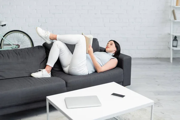 Young pregnant woman reading book on couch near gadgets on table in living room — Stock Photo