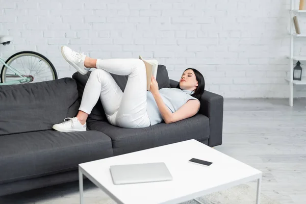 Jovem grávida leitura livro no sofá perto de gadgets na mesa na sala de estar — Fotografia de Stock