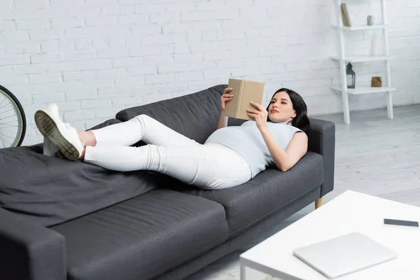 Беременная женщина читает книгу, лежа на диване рядом с размытыми гаджетами на столе в гостиной — стоковое фото