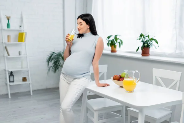 Joven embarazada bebiendo jugo de naranja cerca de manzanas frescas en la mesa de la cocina — Stock Photo