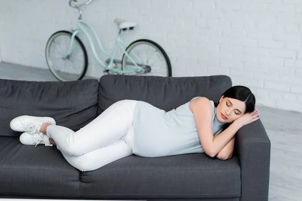 Vue pleine longueur de la jeune femme enceinte dormant sur le canapé près du vélo flou dans le salon — Photo de stock