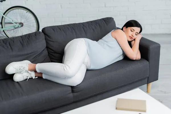 Vista completa de la mujer embarazada morena descansando en el sofá en la sala de estar - foto de stock