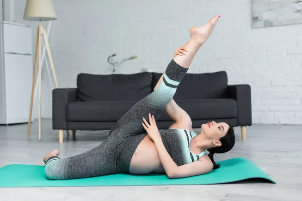 Mujer embarazada joven estirando con la pierna levantada en la estera del yoga en el país - foto de stock