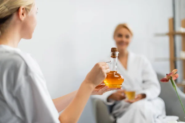Spécialiste du spa blonde tenant cruche avec de l'huile de massage près client flou — Photo de stock