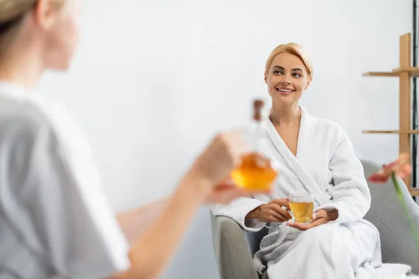 Mujer feliz en albornoz sosteniendo una taza de té mientras mira al especialista en spa borroso con jarra de aceite de masaje - foto de stock