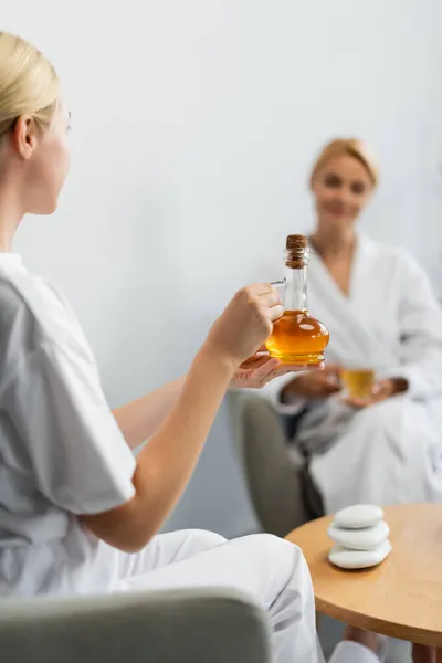 Spécialiste spa blonde tenant cruche en verre avec huile de massage près du client flou — Photo de stock