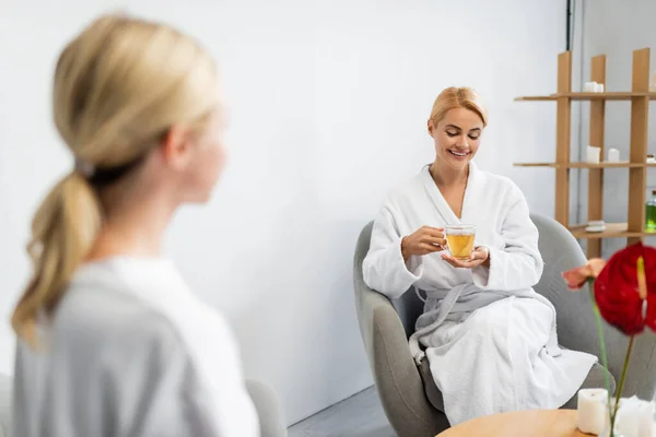 Fröhliche Frau im Bademantel mit einer Tasse Tee im Sitzen in der Nähe eines verschwommenen Wellness-Spezialisten — Stockfoto