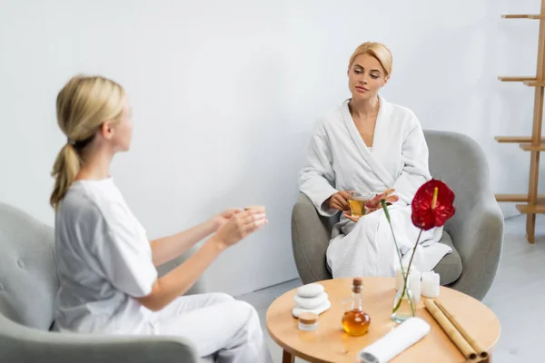 Blonde Kundin im Bademantel mit Tasse Tee beim Anblick eines verschwommenen Wellness-Spezialisten mit Creme-Behälter — Stockfoto