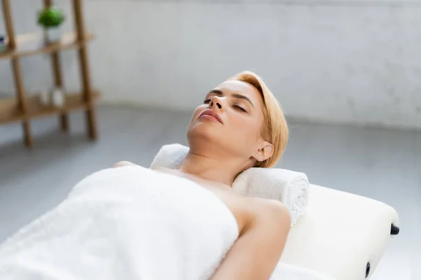 Blonde woman with closed eyes lying on tower roll and massage table in spa center — Stock Photo