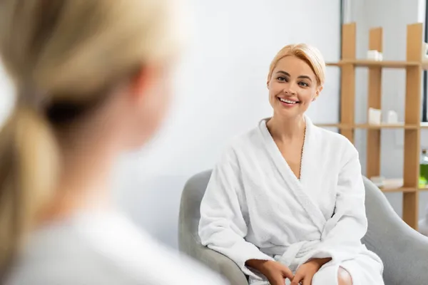 Happy client in bathrobe looking at blurred spa specialist — Stock Photo