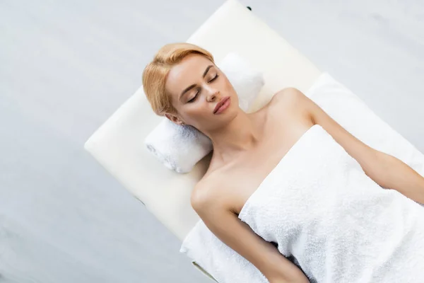 Top view of blonde woman with bare shoulders lying on tower roll and massage table in spa center — Stock Photo