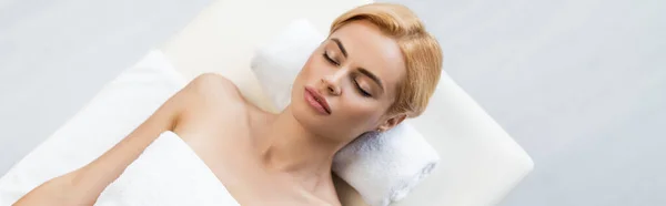 Top view of blonde woman with bare shoulders lying on tower roll and massage table in spa center, banner — Stock Photo