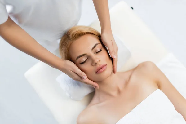 Vista dall'alto del massaggiatore che fa massaggio facciale del cliente biondo con gli occhi chiusi — Foto stock