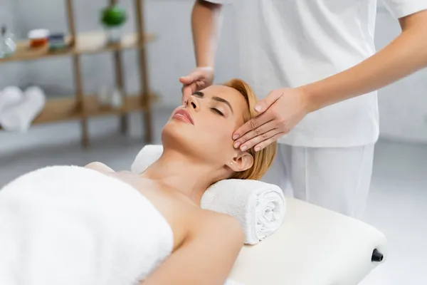 Masajista haciendo masaje de cabeza de cliente rubio con los ojos cerrados - foto de stock