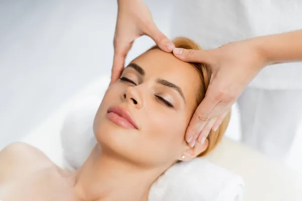 Vue grand angle de masseur faire massage du visage de client blonde avec les yeux fermés — Photo de stock