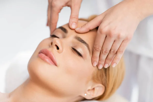 Primer plano del cliente rubio con los ojos cerrados recibiendo masaje facial en el centro de spa - foto de stock