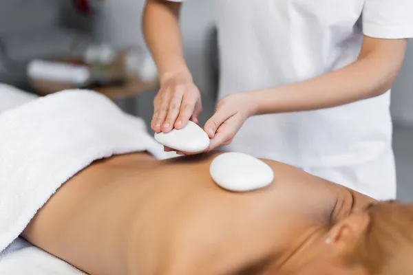 Partial view of blurred masseur placing hot stones on back of client in spa center — Stock Photo