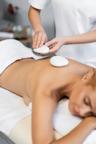 Masseur placing hot stones on back of blurred client in spa center — Stock Photo