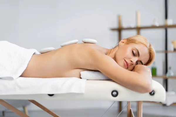 Femme blonde avec les yeux fermés recevant un massage aux pierres chaudes dans le centre spa — Photo de stock