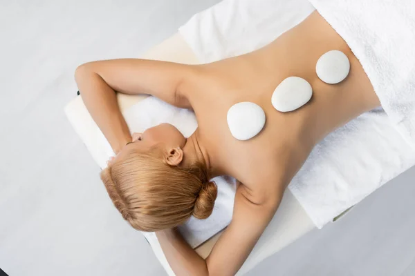 Top view of blonde woman with closed eyes receiving hot stone massage in spa center — Stock Photo