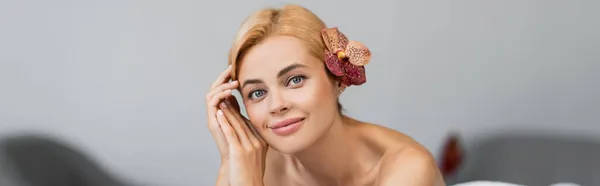Щасливий блондинка клієнт з тропічною квіткою в волоссі дивиться на камеру в спа-центрі, банер — стокове фото