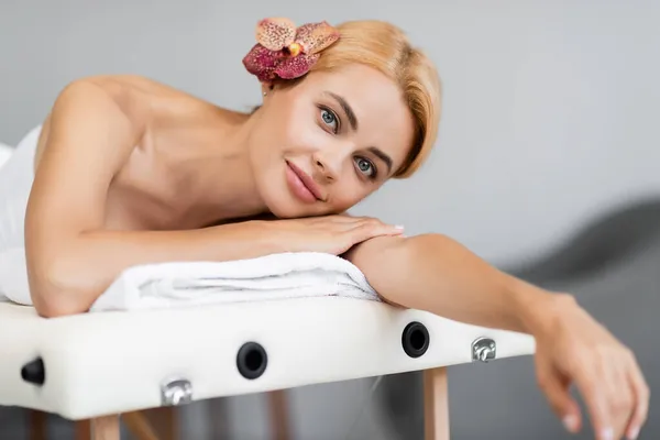Glückliche blonde Frau mit Orchidee im Haar auf Massagetisch liegend — Stockfoto