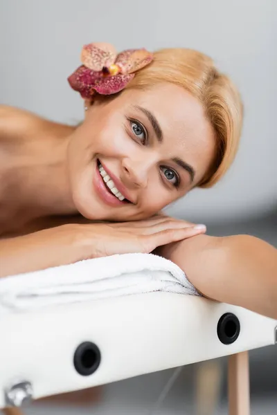Fröhliche blonde Frau mit Orchidee im Haar auf Massagetisch liegend — Stockfoto