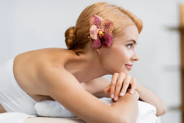 Vue latérale de la femme blonde heureuse avec orchidée dans les cheveux couchés sur la table de massage — Photo de stock
