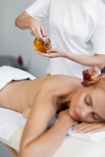 Massaggiatore che tiene la brocca mentre versa olio da massaggio sul retro del cliente offuscato — Foto stock
