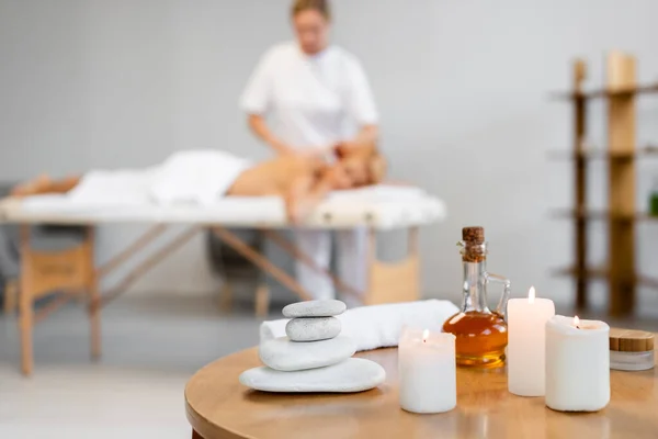 Hot stones near jug with massage oil, candles, and container with cosmetic cream near blurred masseur with client on background — Stock Photo