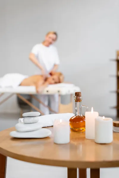 Piedras calientes cerca de aceite de masaje, velas ardientes y rollo de toallas cerca de masajista borroso con el cliente en el fondo — Stock Photo