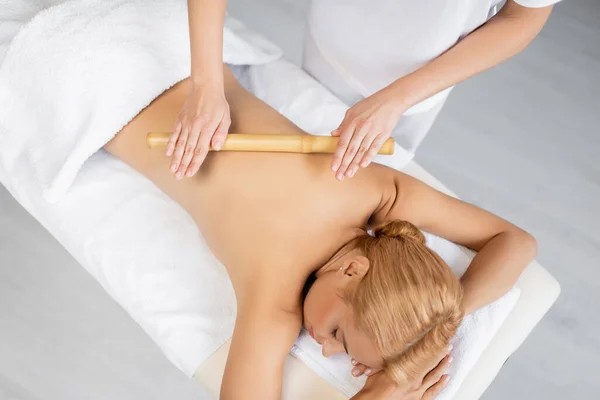 Vista superior de masajista profesional masajeando la espalda de mujer rubia con palo de bambú - foto de stock