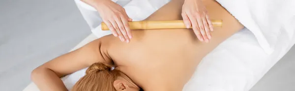 Recortado vista de masajista profesional masaje espalda de mujer rubia con palo de bambú, bandera - foto de stock