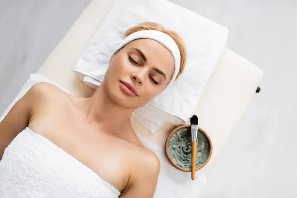 Top view of woman in headband lying on massage table near bowl with clay mask — Stock Photo