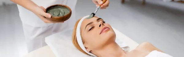 Esthéticienne appliquant un masque d'argile sur le visage de la femme allongée sur une table de massage, bannière — Photo de stock