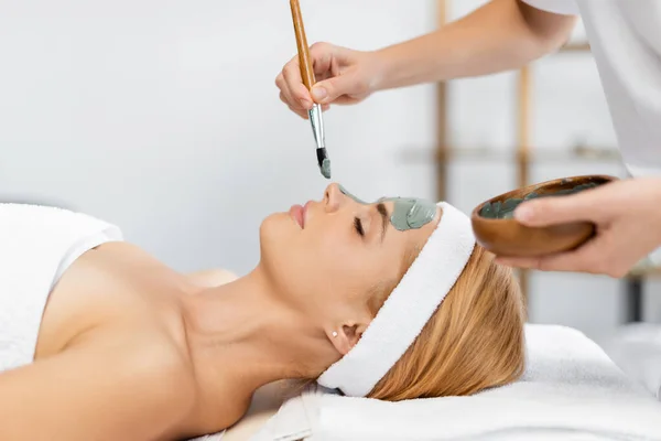 Seitenansicht der Kosmetikerin, die Schale hält und Tonmaske auf das Gesicht der Frau im Wellnesszentrum aufträgt — Stockfoto
